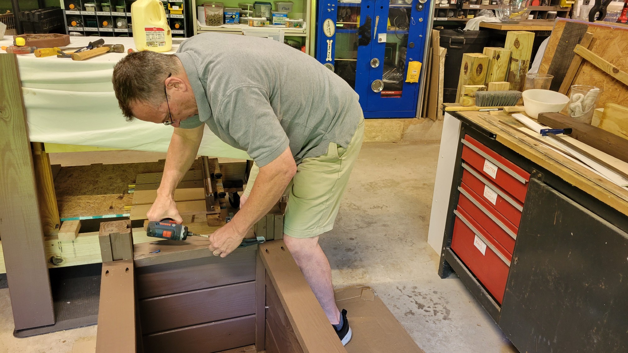 Man in a work garage leaning over a planter that he is building and using a drill
