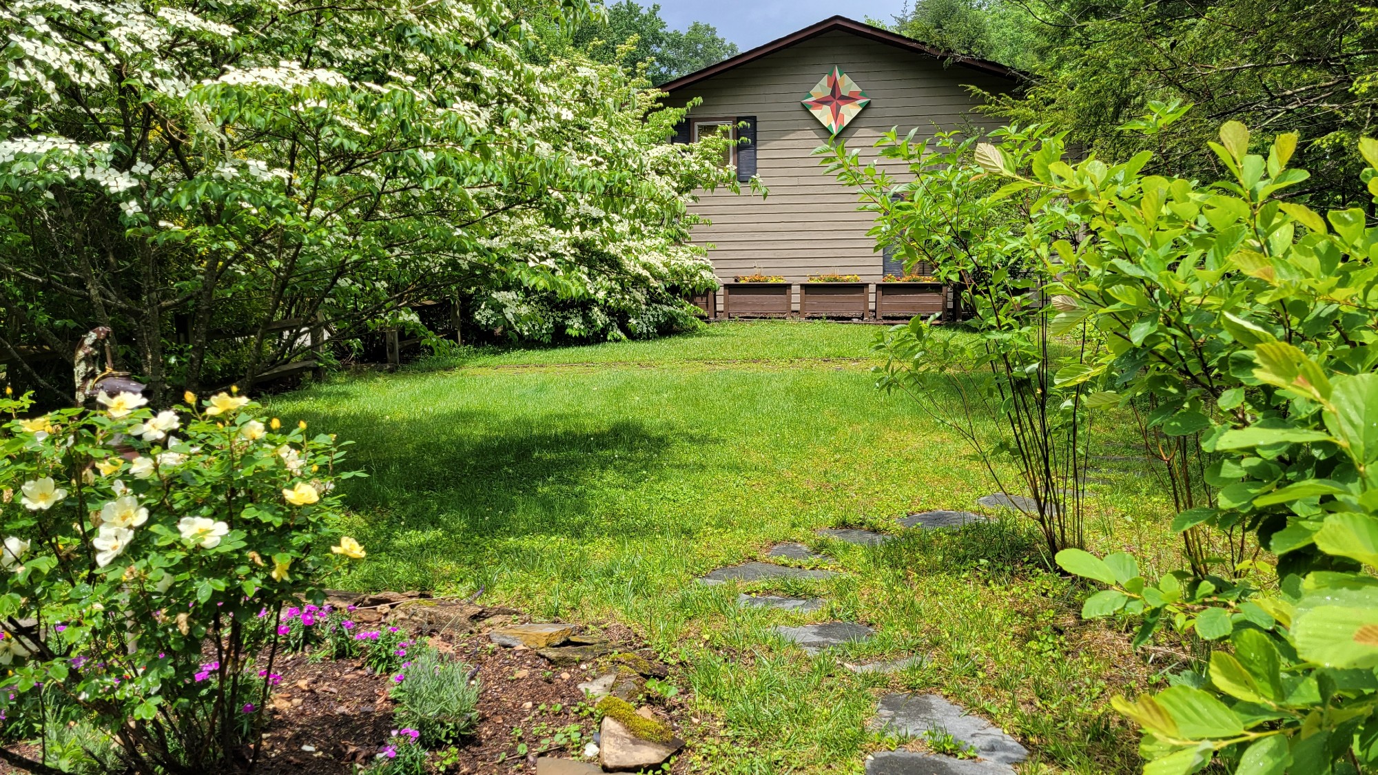 View of a lawn lined with flowers, shrubs and trees with a small house and flower planters in the background