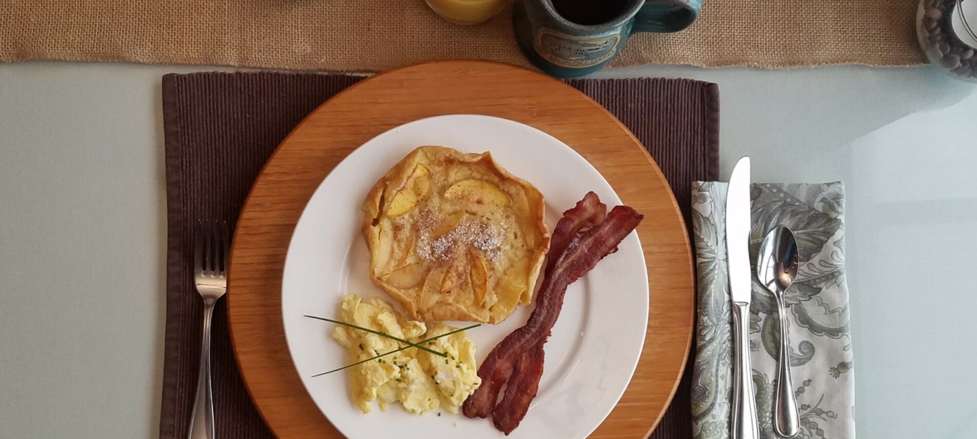 Overhead view of a table with breakfast plate containing peach dutch baby pancake, scrambled eggs and bacon along with coffee in a pottery mug and orange juice