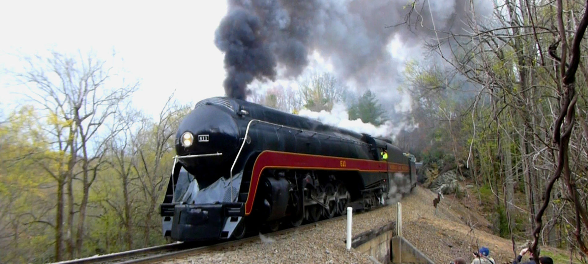 Large train engine with steam coming out of the top as it goes by on railroad tracks