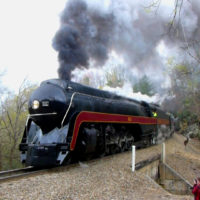 Large train engine with steam coming out of the top as it goes by on railroad tracks