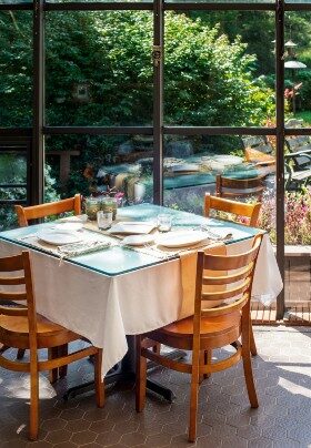 Dining table with four chairs and white tablecloth set in a glassed-in solarium with a garden view