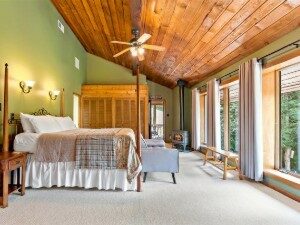 Large bedroom with vaulted cedar plank ceiling, and four poster bed facing wall of floor to ceiling windows