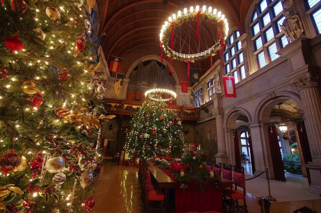 Mansion dining hall with red fabric chairs under round chandeliers and a barrel vaulted ceiling with large decorated Christmas trees on the floor and a pipe organ on the far balcony