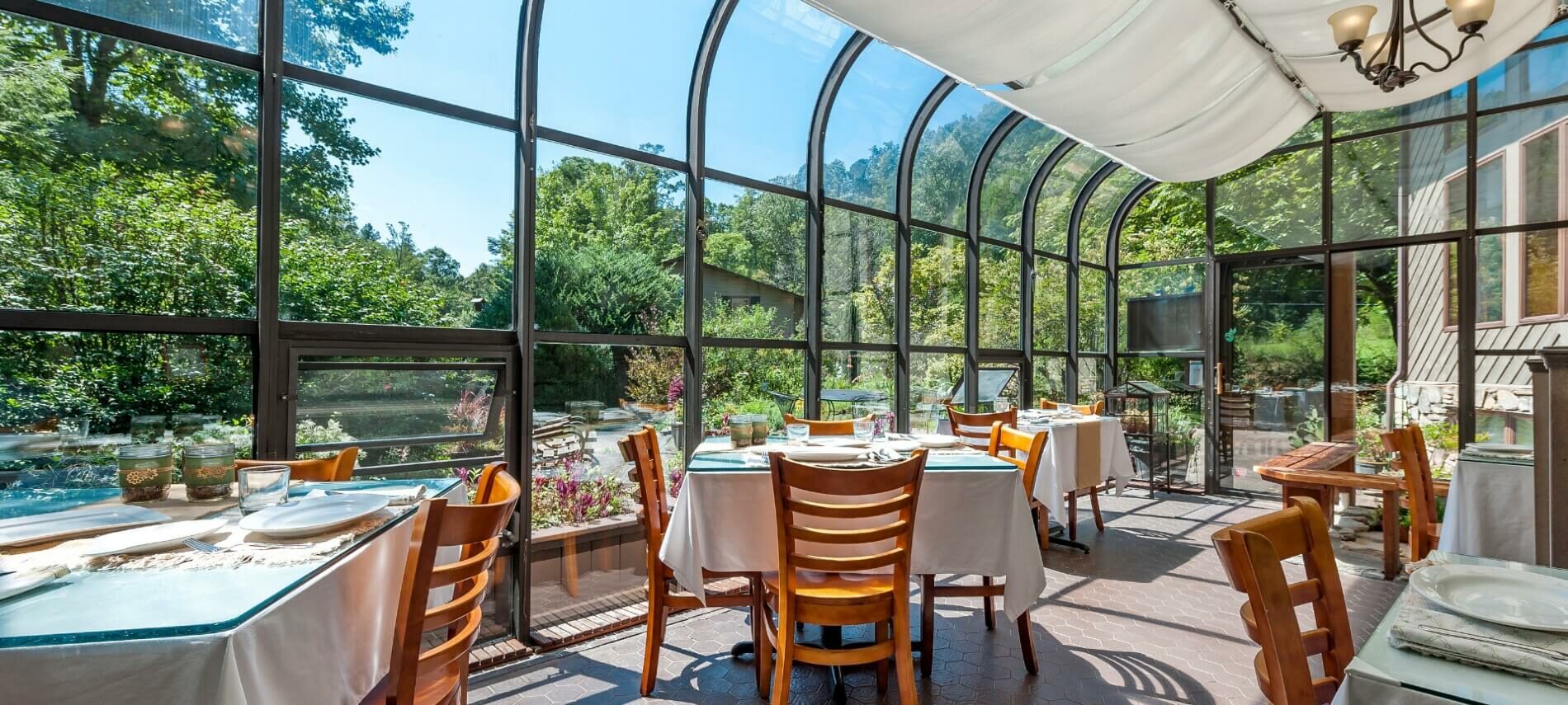 Tables and chairs set for dining in a glassed-in solarium looking out on trees, garden and blue sky