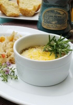 Baked egg dish with cheese and rosemary sprig garnish on plate in front of logo coffee mug and muffins