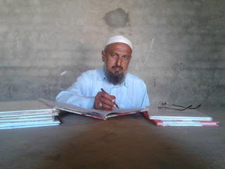 Pakistani school teacher sitting at a table writing in a book