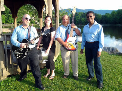 Two men with guitars, a woman with a drum and a fourth band member posing by a lake and gazebo