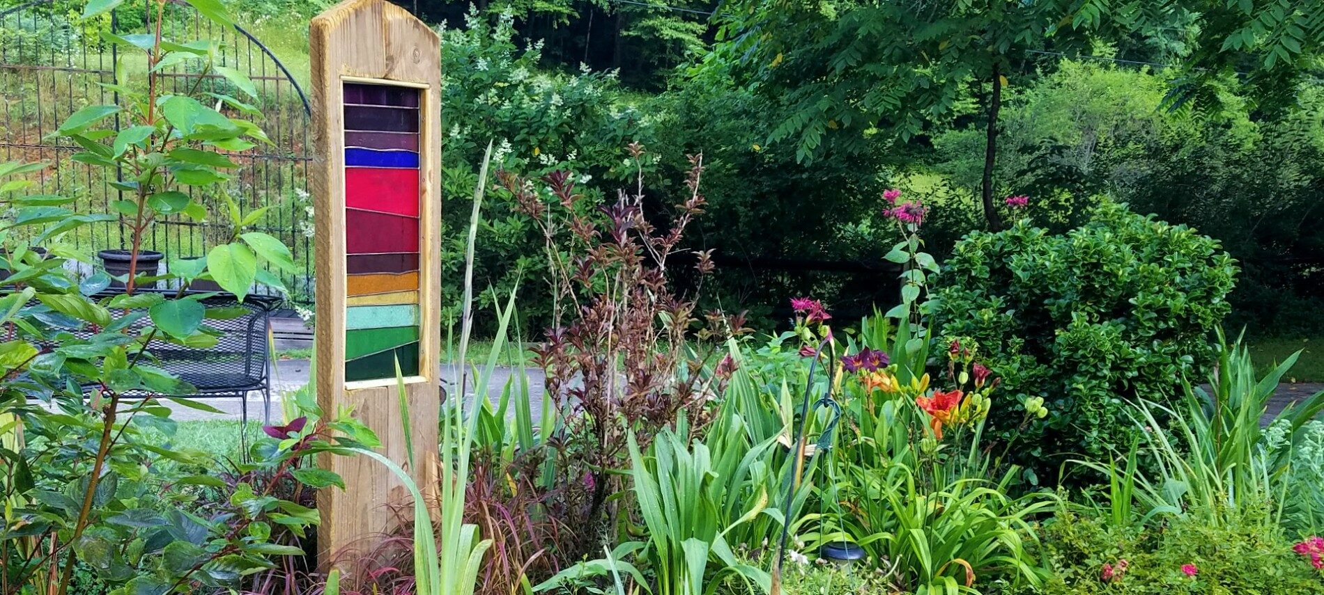 Rectangular Stained Glass Sculpture with layers of colors in a wooden frame set in a garden with flowering trees and a gate in the background